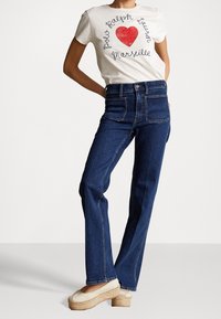 Vit t-shirt med röd hjärtgrafik och svart text, ihop med mörkblå högmidjade jeans och beige plattform espadrillos.