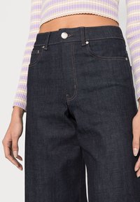 Jeans de ganga escuros com cintura alta, apresentando costura em cobre, cinco bolsos e um fecho de botão prateado na frente.