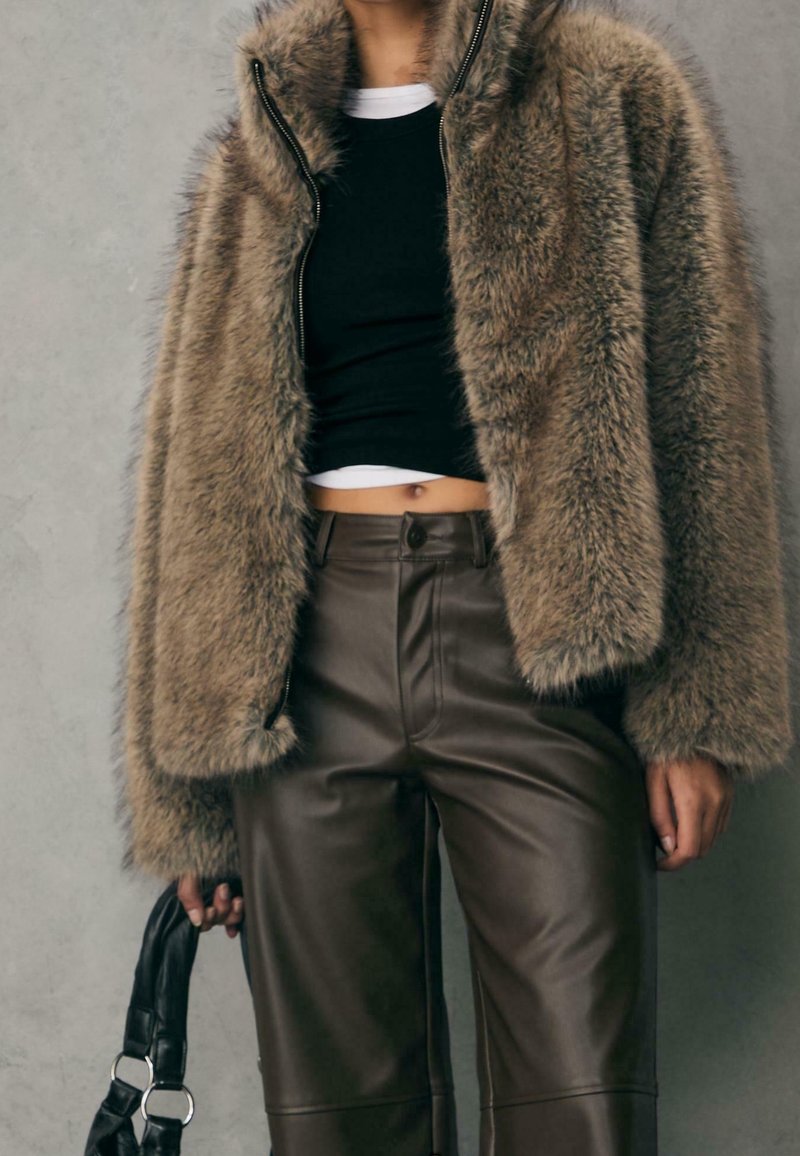 Femme portant une veste en fausse fourrure marron, des hauts superposés courts noir et blanc, un pantalon en cuir marron foncé, tenant un sac à main noir.
