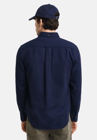 Camicia a maniche lunghe blu navy con piega sul retro, polsini a bottoni e colletto, realizzata in tessuto testurizzato. Il cappello completa l'outfit.