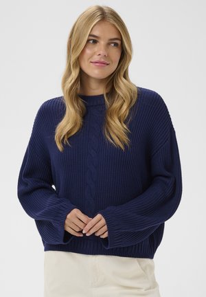 Blondína s vlnitými vlasmi v námornícko modrom pulóvri s vrkočovým vzorom a svetlobéžových nohaviciach, ruky voľne spojené v oblasti pása.