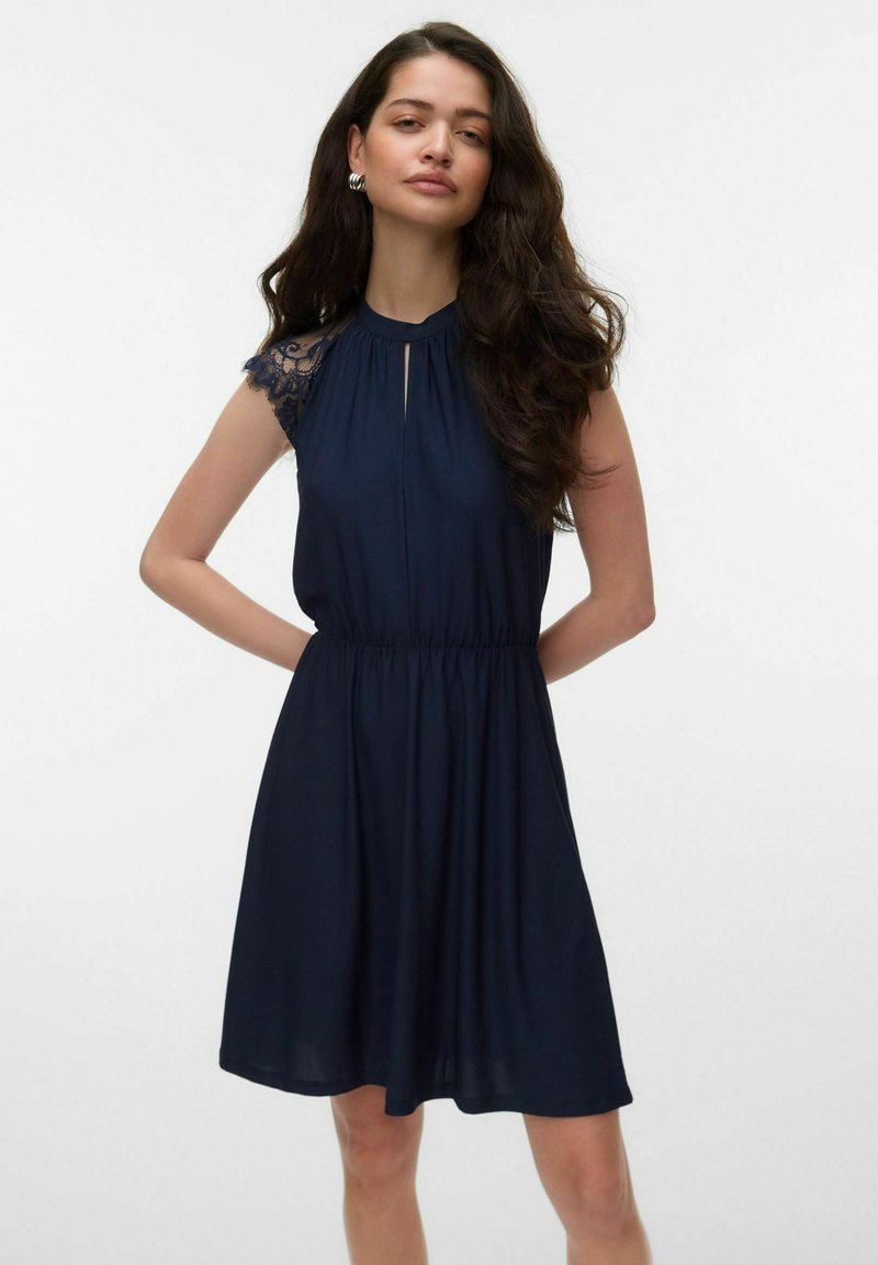 Femme aux longs cheveux noirs portant une robe bleu marine sans manches avec des détails en dentelle aux épaules, debout les mains derrière le dos sur fond blanc.