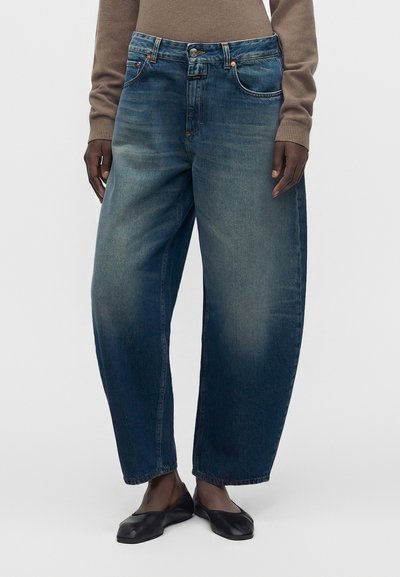 Jeans en denim bleu à jambes larges portés avec des ballerines noires et un top marron à manches longues, montrant le devant et le bas du corps.