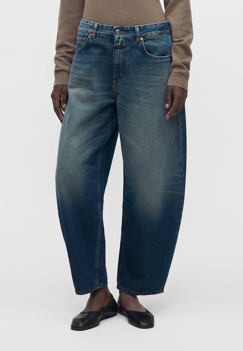Jeans en denim bleu à jambes larges portés avec des ballerines noires et un top marron à manches longues, montrant le devant et le bas du corps.