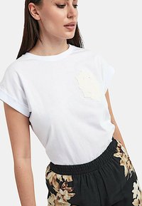 T-shirt in cotone bianco con maniche arrotolate, caratterizzata da un'applicazione floreale bianca testurizzata sul petto. Abbinata a pantaloni neri con motivo floreale.