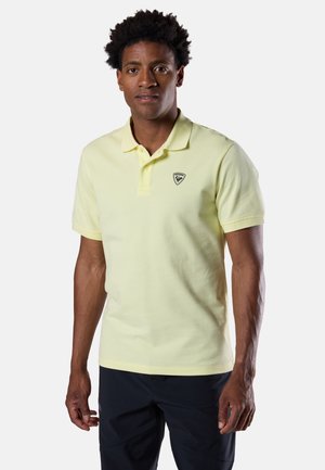 Mann trägt ein hellgelbes Polohemd mit kurzen Ärmeln und Logo auf der Brust sowie eine dunkle Hose, steht vor hellem, einfarbigem Hintergrund.