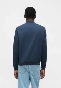 Mann von hinten gesehen, der eine marineblaue Bomberjacke mit geripptem Kragen und hellblauen Jeans trägt, vor einem schlichten weißen Hintergrund.