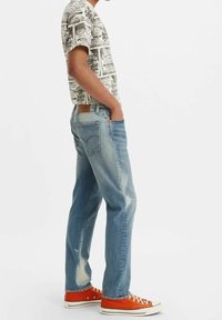 Pantalones vaqueros de mezclilla azul claro con un ajuste relajado, con efecto desgastado y detalles de desgaste natural. Combinados con una camisa estampada y zapatillas naranjas.