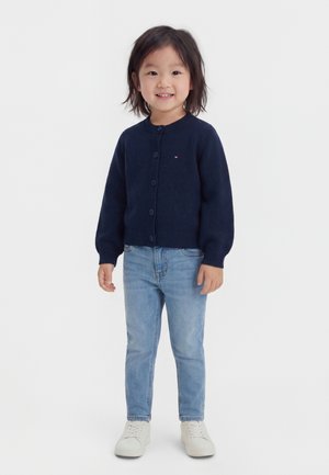 Child standing and smiling, wearing a navy blue buttoned cardigan, light blue jeans, and white sneakers against a plain background.