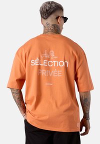 T-shirt en coton orange avec une coupe décontractée. Il présente à l'arrière un texte blanc indiquant "LA Partie Deux Sélection Privée". Manches courtes, col rond.