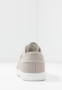 Beige canvas sneaker with a white textured rubber sole, featuring a simple back design and visible stitching details.