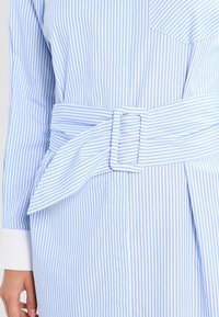 Helles blau-weiß gestreiftes Kleid mit einem Taillengürtel, breitem Kragen, langen Ärmeln und einer Brusttasche.