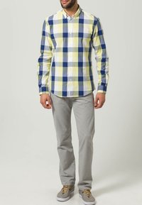 Camicia a quadri gialla, blu e bianca, realizzata in cotone, con maniche lunghe e chiusura a bottoni; abbinata a pantaloni grigi e scarpe chiare.
