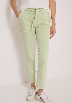 Personne portant un pantalon décontracté vert clair avec des poches plaquées à l’avant et des baskets blanches avec des accents rouges sur un fond uni.