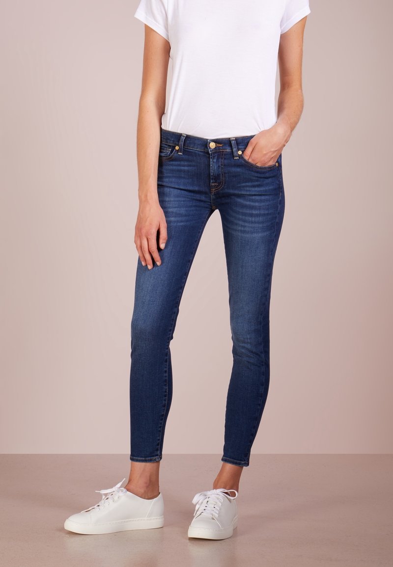 Rikkaita tummansinisistä farkuista, joissa on keskimäinen vyötärö, viisi taskua ja kultaiset korostukset, yhdistetty valkoisiin tennareihin ja yksinkertaiseen valkoiseen t-paitaan.