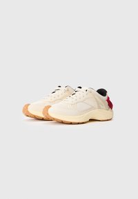 Zapatillas deportivas color crema con paneles de malla y ante, con suela de goma, detalles negros en la lengüeta y un detalle rojo en el talón.