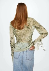 Crop top a maniche lunghe, trasparente, con stampa animale verde e beige e dettagli con fiocchi ai polsi, indossato sopra una minigonna in denim azzurro.