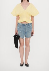 Gelbes, kurz geschnittenes Oberteil mit V-Ausschnitt und plissierten Ärmeln, kombiniert mit zerrissenen Jeansshorts und einer schwarzen geflochtenen Tasche. Schwarze Ballerinas runden den Look ab.