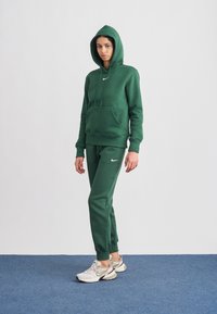 Camisola verde com capuz, bolso frontal e cordão, combinada com calças de fato de treino correspondentes. Ambos apresentam um logótipo branco da Nike. A modelo usa ténis bege.