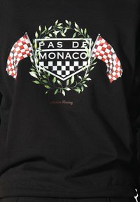 Tessuto nero con un motivo di bandiere a scacchi rosse e bianche, una corona di alloro verde e il testo "PAS DE MONACO" e "Atelier Racing."