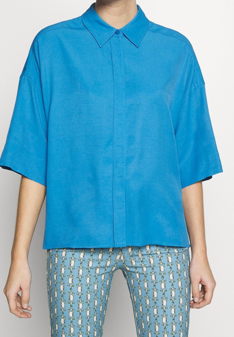 Korte mouwen, button-up shirt van heldere blauwe stof. Heeft een klassieke kraag en een comfortabele pasvorm, gecombineerd met gestreepte blauwe broek.