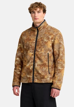 Giovane uomo che indossa una giacca in pile marrone e beige a fantasia con cerniera nera e pantaloni neri su sfondo bianco.