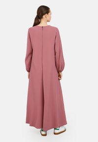 Dlouhé, růžové šaty vyrobené z hladkého materiálu s vysokým výstřihem, nabíranými dlouhými rukávy a zipem na zádech. Stylizované s bílými a zelenými teniskami.