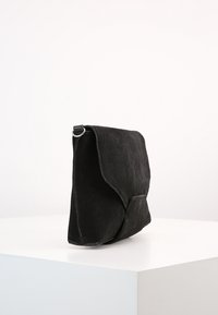 Černá semišová taška přes rameno s klopou, vybavená jedním stříbrným kroužkem na ramenním popruhu a texturovaným povrchem.
