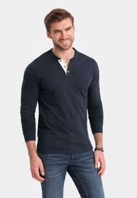 Maglietta henley a maniche lunghe blu navy con una placca di bottoni bianchi e bottoni marroni. Indossata con jeans blu, presenta una vestibilità comoda e una texture liscia.