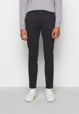 Hombre con pantalones negros de ajuste slim y zapatillas blancas, de pie contra un fondo claro y liso.