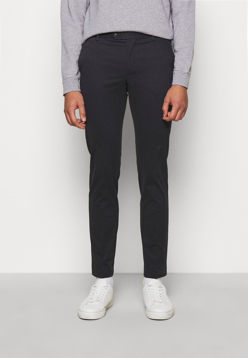 Homme portant un pantalon noir slim et des baskets blanches, debout devant un fond clair uni.