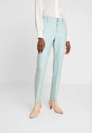 Femme portant un pantalon bleu clair sur mesure, une chemise blanche à manches longues et des bottines couleur nude, debout sur un fond clair uni.