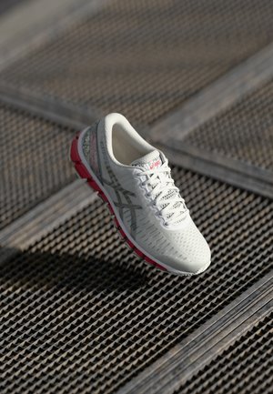 Witte en grijze Asics hardloopschoen met roze zool details, rustend op een metalen rooster buiten.