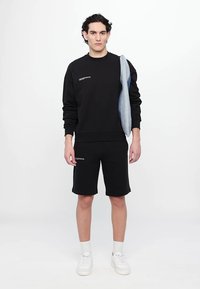 Schwarzer Sweatshirt mit leicht gerippter Textur, Logodetail auf der Brust, kombiniert mit schwarzen Shorts. Weiße Sneakers und eine blaue Jeansjacke über der Schulter.