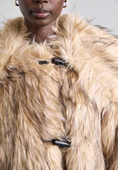 Fauxfur jas in lichtbeige met donkere strepen, met een fluffy textuur en zwarte togglesluitingen aan de voorkant.