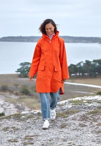 Bright orange raincoat with a hood, two front pockets, and a high-low hem, paired with light blue jeans and white sneakers.