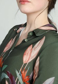 Blusa verde scuro con stampa floreale caratterizzata da grandi foglie in tonalità di rosa, arancione e teal; colletto con bottoni a pressione delicati.