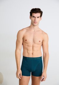 Τιρκουάζ σλιπ με απαλή υφή και λάστιχο με λογότυπο. Το μοντέλο στέκεται σε ένα ουδέτερο φόντο, παρουσιάζοντας την εφαρμογή και τον σχεδιασμό.