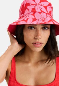 Červený klobúk s širokým okrajom a štruktúrovaným tvarom, pokrytý ružovým kvetinovým vzorom a logom Roxy. Tkanina vyzerá textúrovaná.