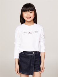 Camicia bianca a maniche lunghe con logo "TOMMY HILFIGER", abbinata a una gonna a portafoglio in denim scuro con bottoni frontali e dettagli di cucitura.