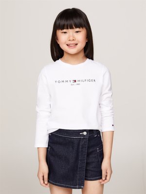 Camicia bianca a maniche lunghe con logo "TOMMY HILFIGER", abbinata a una gonna a portafoglio in denim scuro con bottoni frontali e dettagli di cucitura.