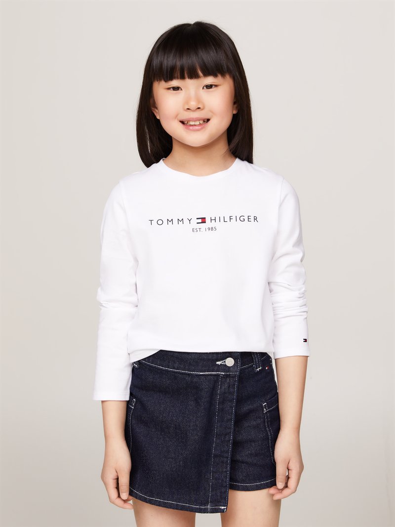Camicia bianca a maniche lunghe con logo "TOMMY HILFIGER", abbinata a una gonna a portafoglio in denim scuro con bottoni frontali e dettagli di cucitura.