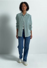 Camicia button-up in corduroy color turchese chiaro con colletto, abbinata a jeans blu a gamba dritta e sneakers bianche. Sfondo da studio.