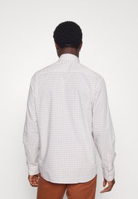 Homme aux cheveux courts et bouclés portant une chemise à manches longues à carreaux blancs et beiges et un pantalon couleur rouille, vu de dos.