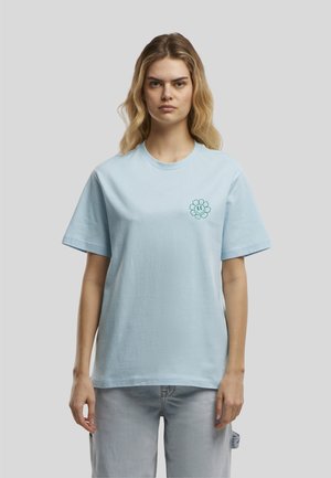 Jeune femme aux cheveux blonds ondulés portant un T-shirt bleu clair avec un petit motif fleur sourire sur le côté gauche de la poitrine.