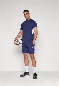 Laadukkaat tummansiniset urheiluvaatteet valkoisilla yksityiskohdilla, yhdistettynä sopiviin shortseihin. Malli pitää jalkapalloa käsissään, jalassa mustat urheilukengät ja valkoiset sukat.