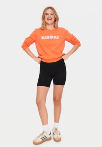 Oranje sweatshirt met "BISOUS" in het wit, gecombineerd met zwarte fietsbroeken. Witte enkelkousen en sneakers met beige, groene en crèmekleurige accenten.