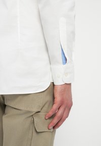 Hvit skjorte med lange ermer, med blå detaljer ved mansjettene. Skjorten er kombinert med beige cargo-bukser som viser lommedetaljer.