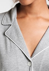 Camisola de pijama cinza em algodão com gola em bico, com acabamento em branco nas bordas e um único botão no peito. Textura suave e design ajustado.