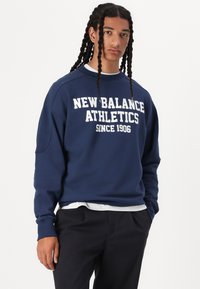 Kék pulóver 100% pamutból, klasszikus kerek nyakkal. Az "New Balance Athletics" felirat díszíti, és laza fazonú. Fekete nadrággal párosítva.